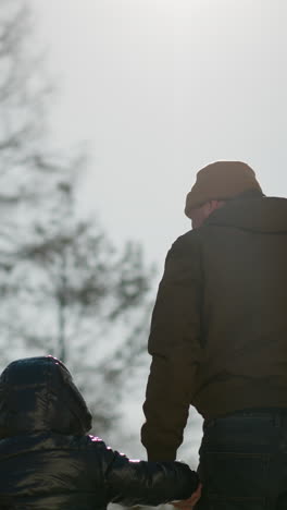 아버지와 아들은 야외에서 손을 잡고 고, 아버지가 는 동안 오른손을 재 주머니에 넣고, 나무와 밝은 하늘을 볼 수 있습니다.