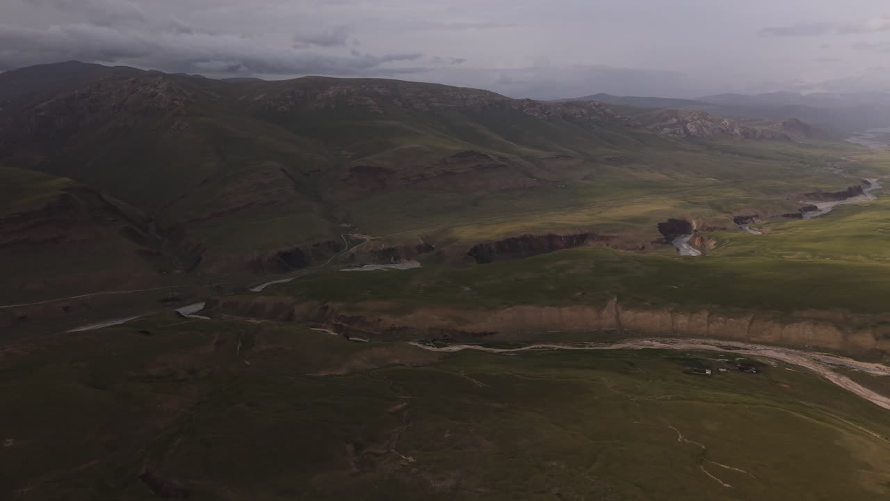 Kyrgyzstan - A Winding River Cuts through Wide Green Valleys Framed by Rugged, Shadowed Mountains Under a Cloudy Sky - Aerial Drone Shot