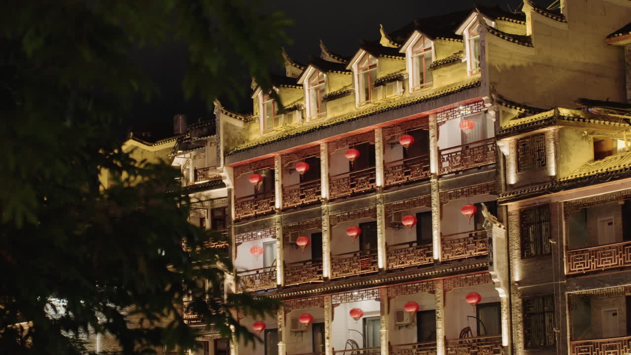 Pousada chinesa iluminada à noite, varandas ornamentadas alinhadas com lanternas vermelhas brilhando através da folhagem das árvores, iluminação exterior dourada e quente destaca beirais tradicionais e colunas decorativas