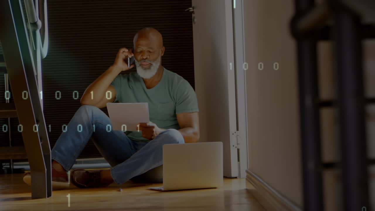 Animation of binary coding over african american man holding document talking on smartphone at home
