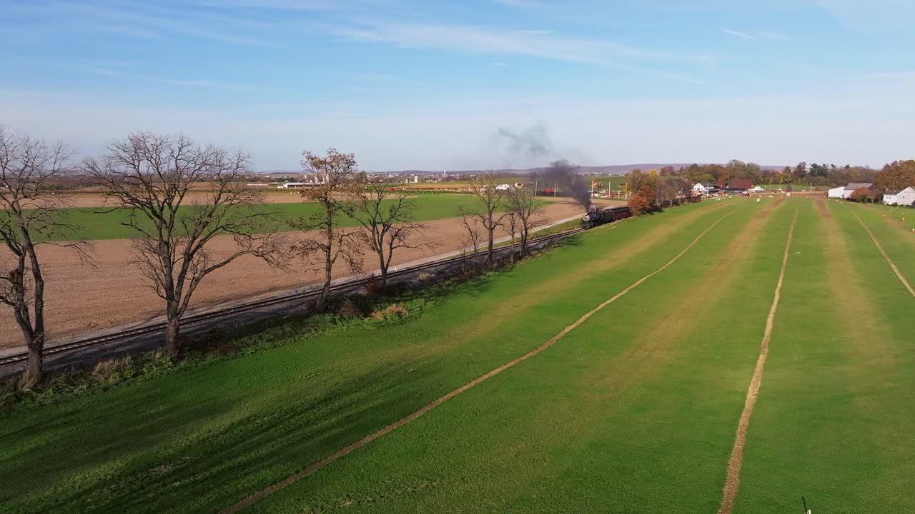 una vista aérea de una sola vía férrea que atraviesa tierras de cultivo mientras un tren de vapor se acerca en la distancia en un otoño soleado