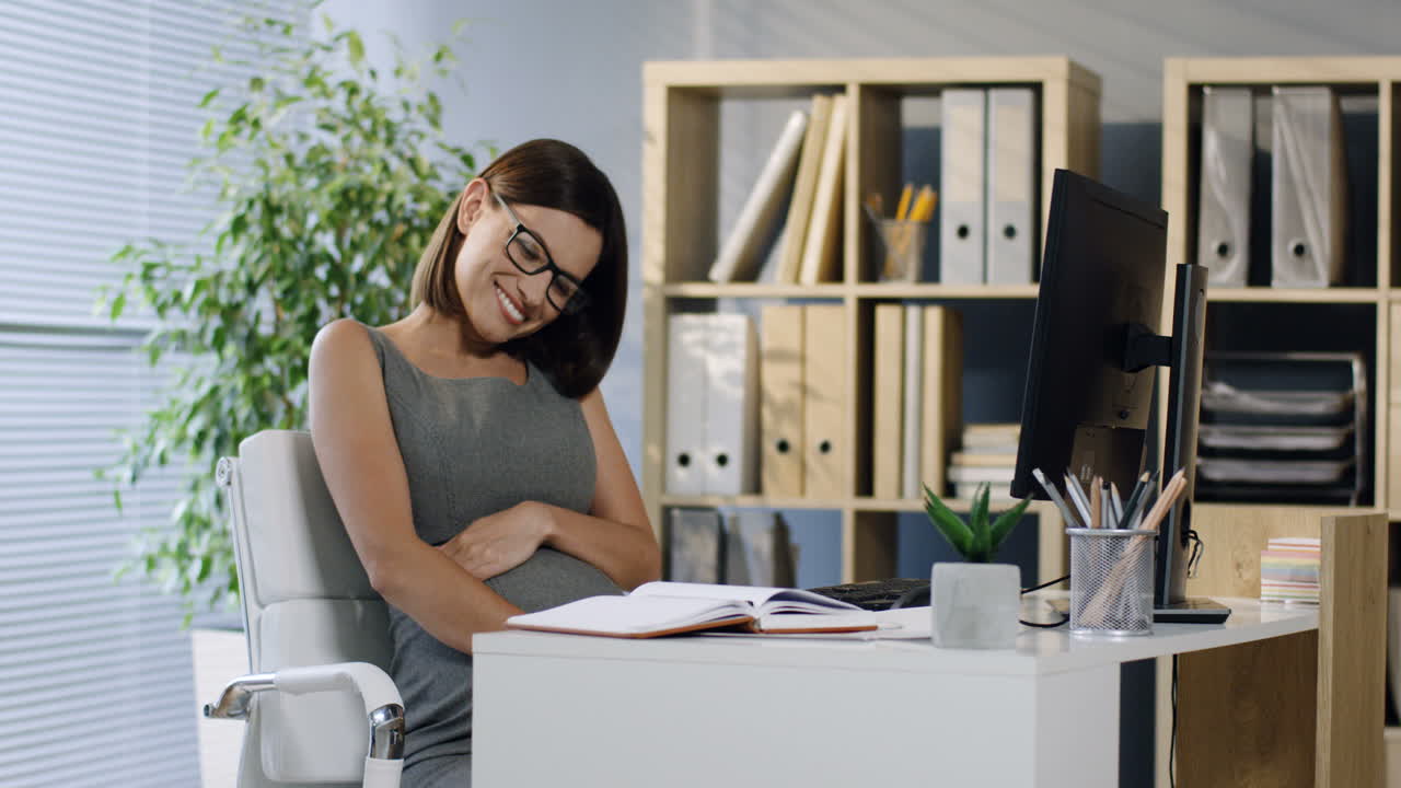 Pregnant Smiling Businesswoman In Glasses Sitting In The Office While Caressing Her Belly
