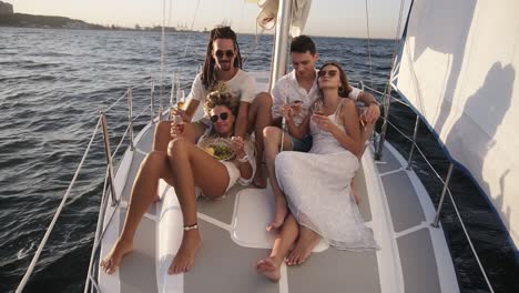 A-company-of-young-happy-friends-or-two-couples-sail-on-a-yacht-bow-on-a-summer-day,-friends,-men-and-women-relax-in-the-sun,-drinking-champagne-and-eating-fruits.-Beautiful,-mild-sunlight-and-sea-on-the-background.-Slow-motion