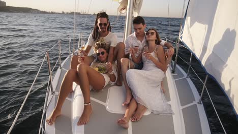 una compañía de jóvenes amigos felices o dos parejas navegan en una proa de yate en un día de verano, amigos, hombres y mujeres se relajan al sol, bebiendo champán y comiendo frutas. hermosa, suave luz solar y mar en el fondo. cámara lenta