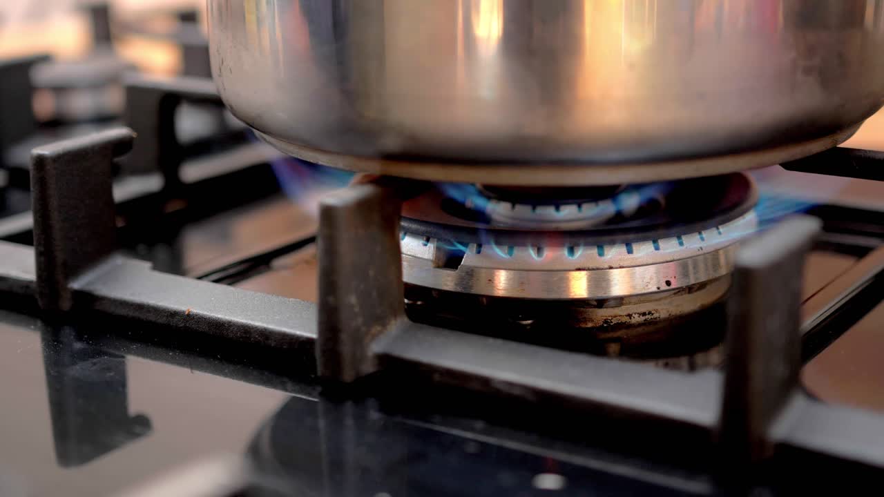 Fornello a gas acceso con fiamme blu che riscaldano una pentola in acciaio inossidabile in una cucina. La scena mostra l'elemento del fornello che fornisce calore per la cottura. Primo piano.