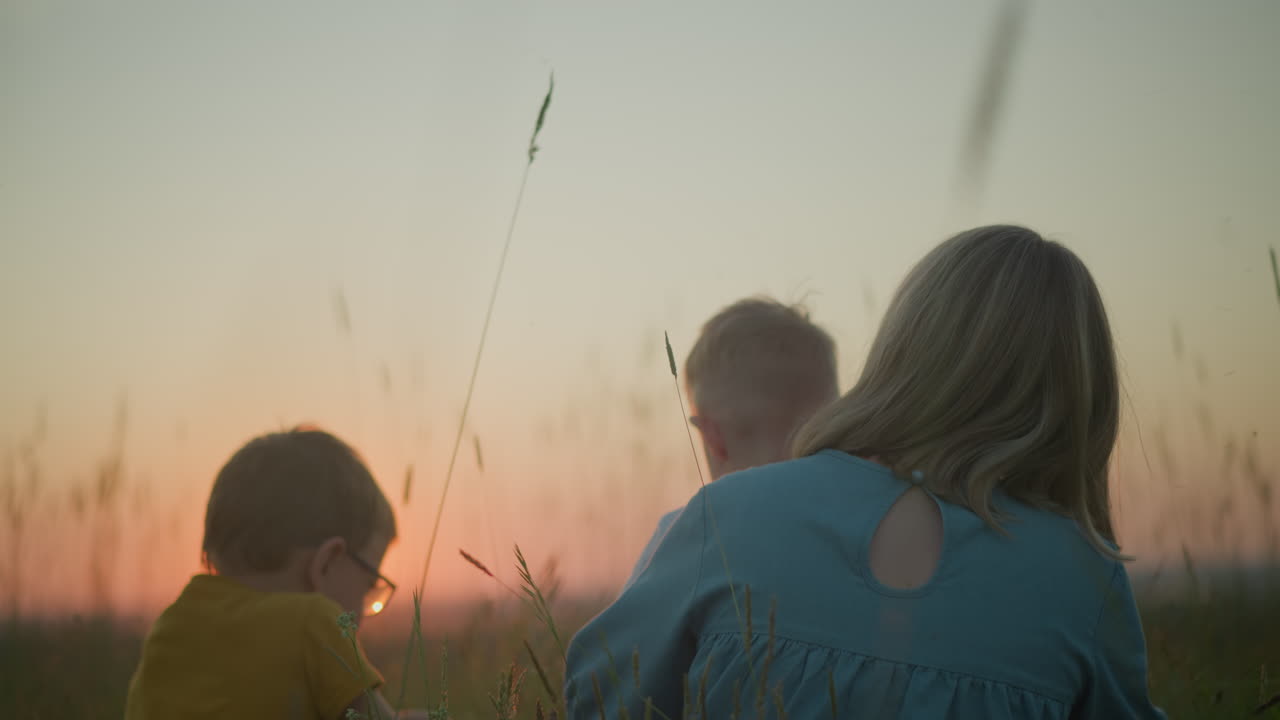 Back view of a mother in a blue gown sitting in a field with her two children, watching the sunset over a tranquil lake. The younger child is sitting on her lap, while the older child, sits beside her