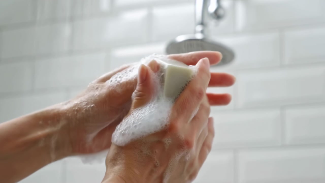 Primer plano de manos lavando una esponja con jabón debajo de un grifo de baño, con un fondo de azulejos blancos y luz lateral suave en un entorno interior sencillo.