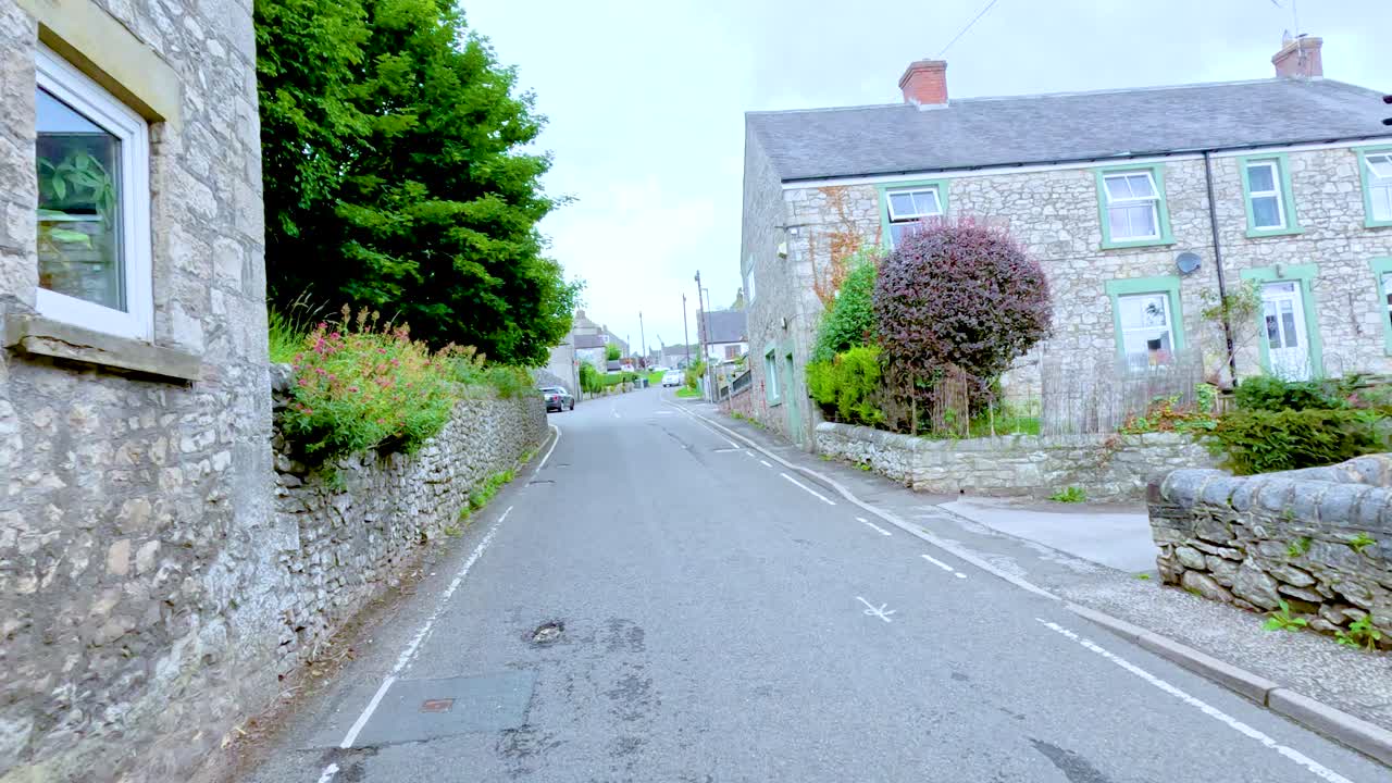 A vehicle travels along a narrow residential street lined with historic stone houses in Buxton, England, under soft daylight with smooth camera movement