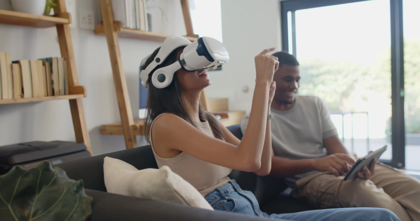 Couple at home, woman putting on VR headset, gesturing to control VR, man tapping tablet