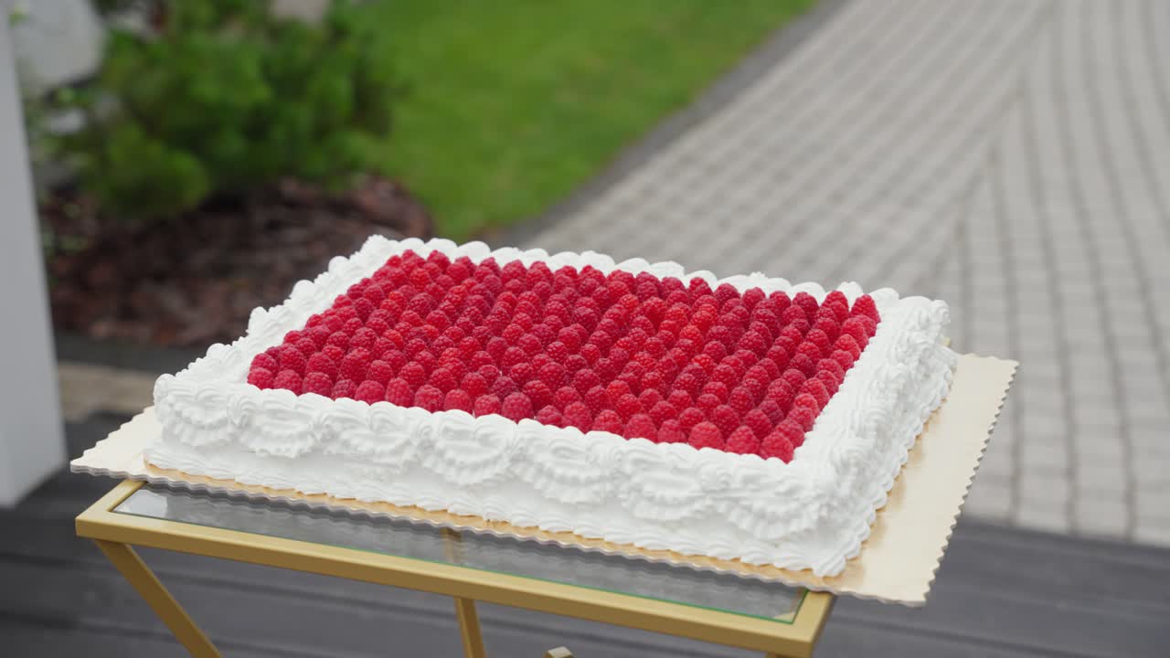 A beautifully decorated rectangular raspberry cake with white cream icing, placed on a glass and gold stand outdoors, perfect for a celebration or wedding