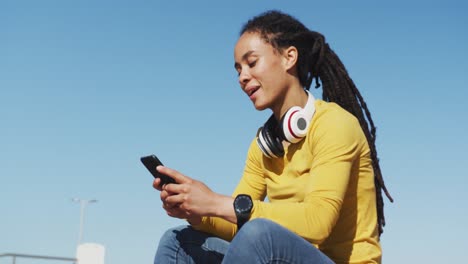 African-american-woman-sitting-using-smartphone-on-promenade-by-the-sea