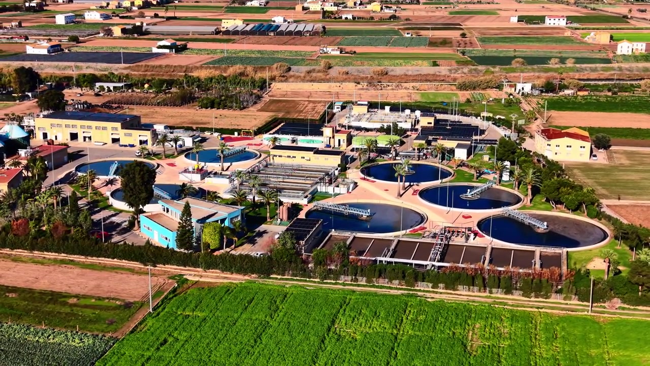 Aerial view of wastewater treatment plant with circular clarifier amid farmland, Valencia, Spain. Blue basin, palm tree, solar panel array and yellow building surrounded by green crop.