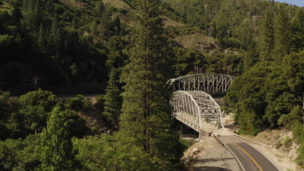 Forward rising aerial, Plumas National Forest above highway 70 with two through arch bridges crossing river in valley, sunny day, passing large pine tree