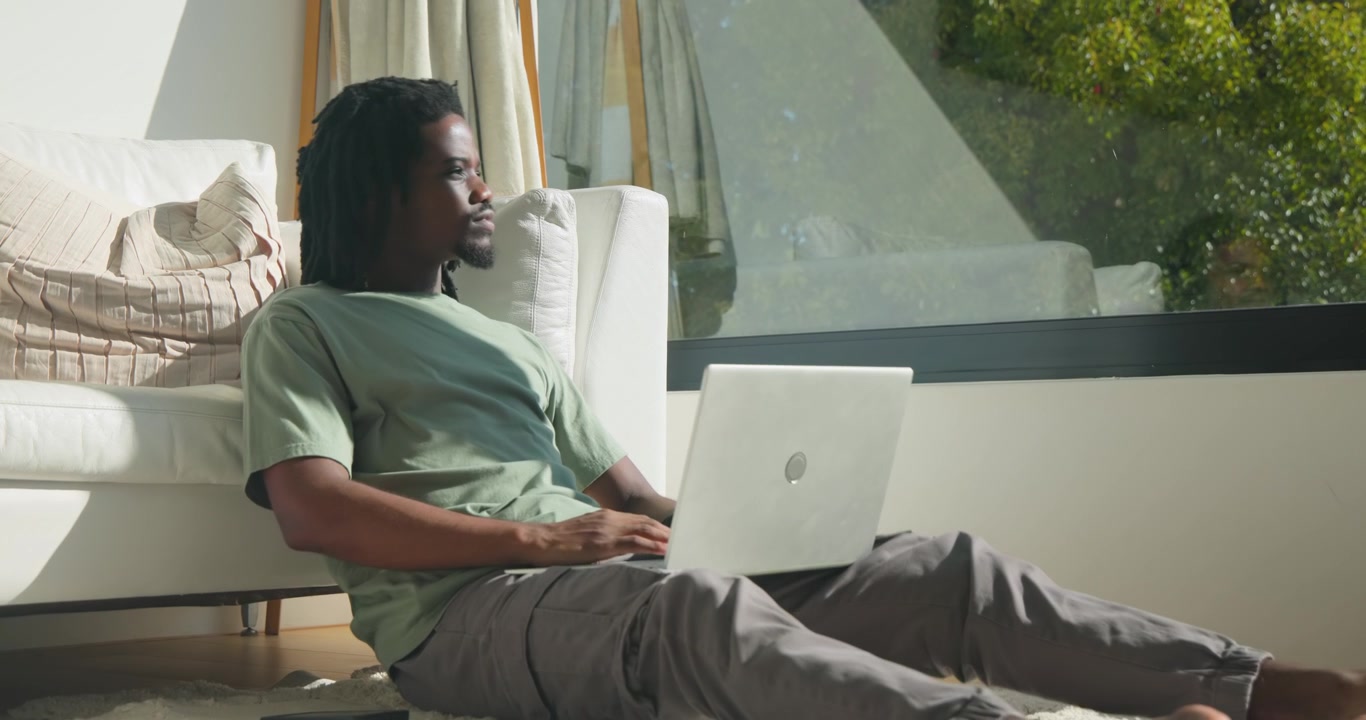 African man opening laptop typing, studying on floor leaning against sofa by window, gazing outside