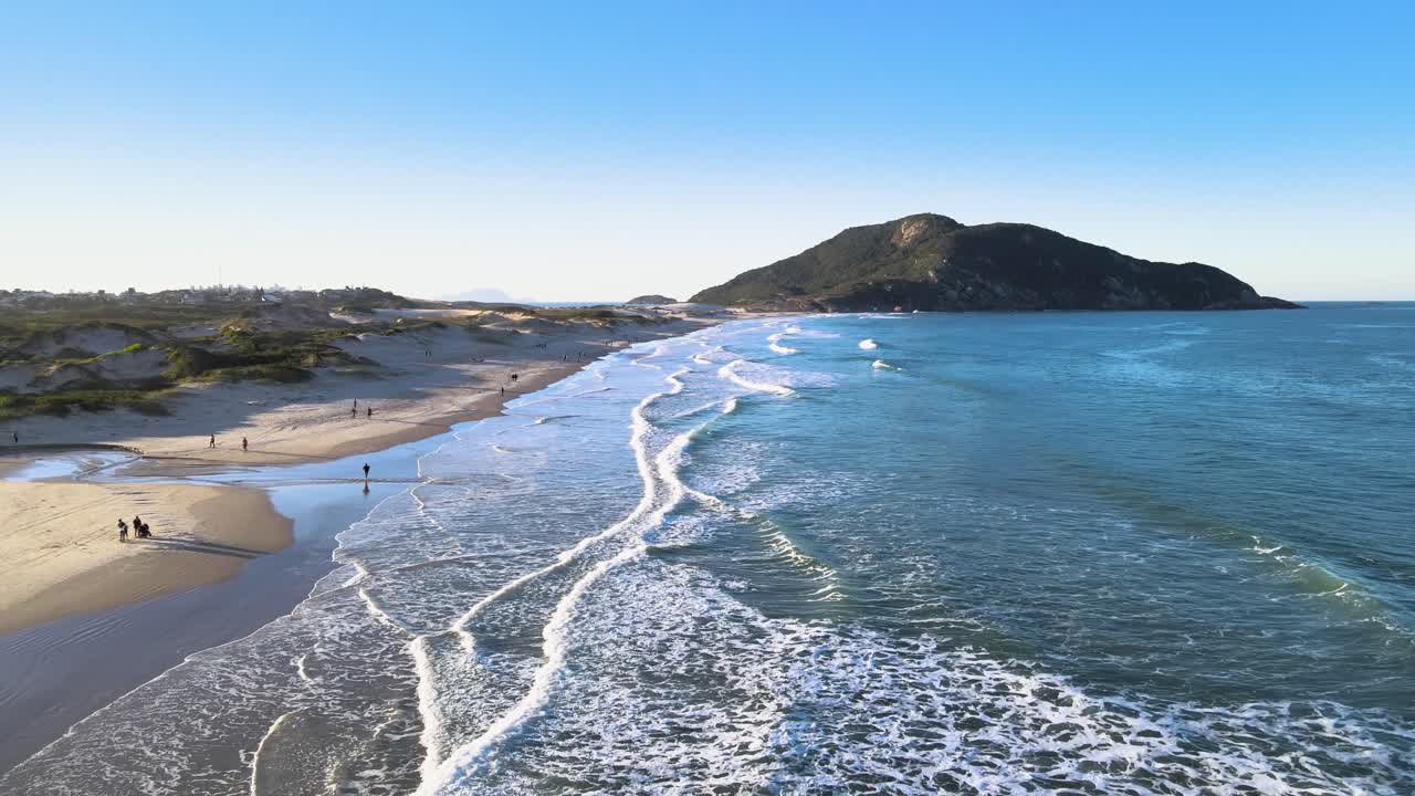 Ripresa aerea cinematografica in allontanamento della spiaggia di Santinho a Florianopolis, Brasile, che rivela onde oceaniche turchesi, dune di sabbia bianca e il monte Morro dos Ingleses sotto un cielo azzurro e limpido