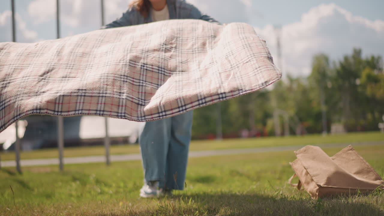 Kvinde knæler og organiserer hyggelig picnicopsætning. Kvinde forbereder afslappet udendørs picnic på solbeskinnet græsområde. Kaukasisk kvinde knæler for at arrangere tæppe og ejendele i naturen