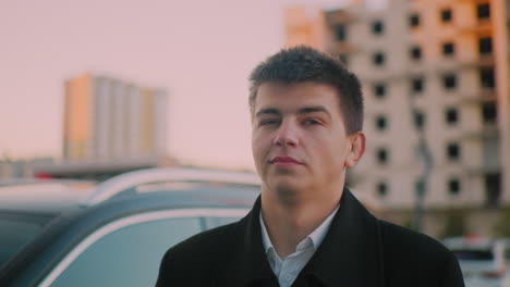 investor standing close to car with subtle smile, background shows distant building at dusk, portraying confident business professional enjoying success in an urban environment