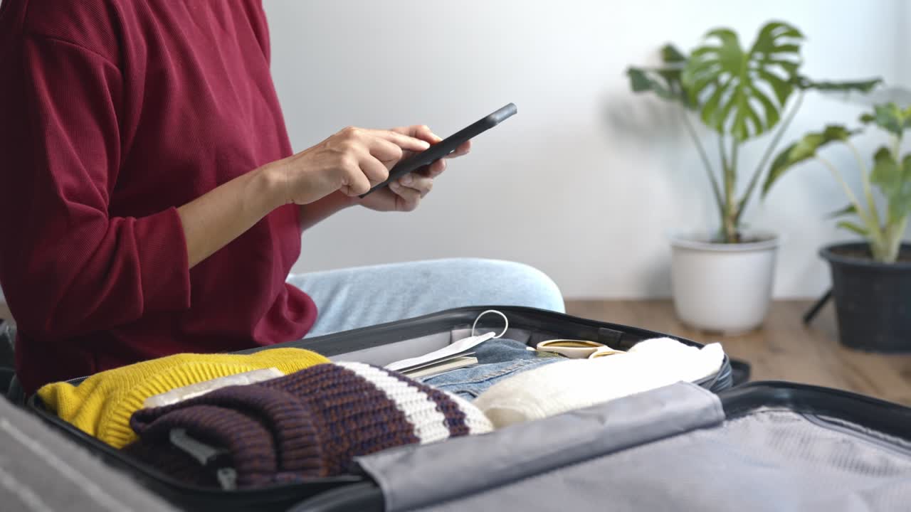 woman using smartphone checklist getting ready for travel, packing clothes and accessories in suitcase. Concept of traveling, adventure.