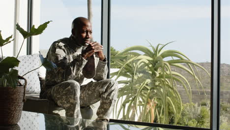 sitting in military uniform, african american man contemplating by large window with plants