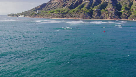 Mientras-El-Dron-Se-Aleja-De-La-Orilla-De-Diamond-Head-En-La-Isla-De-Oahu,-Los-Surfistas-Y-Practicantes-De-Kitesurf-Disfrutan-Deslizándose-Sobre-La-Superficie-De-Las-Aguas-Turquesas-En-Hawai.