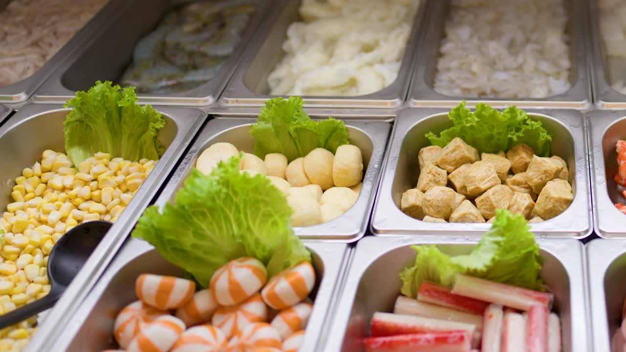 Camera pans across trays of noodles, vegetables, and proteins under bright buffet lighting