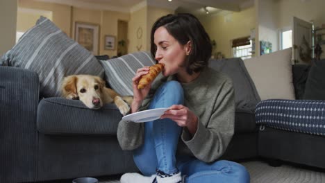 Lächelnde-Kaukasische-Frau,-Die-Ein-Croissant-Isst-Und-Ihren-Hund-Auf-Dem-Sofa-Neben-Ihr-Streichelt