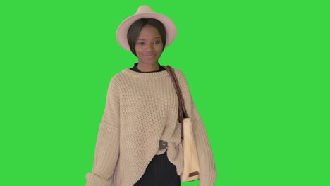 happy african american woman in hat and knitwear walking with shopping bags on a green screen, chroma key