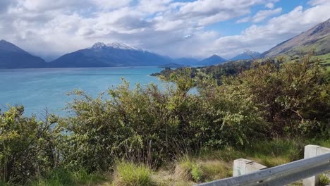 Panning-view-of-road-and-snow-capped-mountains-and-dramatic-light-shining-down-on-the-turquoise-lake-on-the-road-from-Queenstown-to-Glenorchy