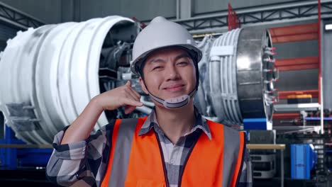 close up of asian male engineer with safety helmet smiling to camera and making call me gesture while standing with airplane engine maintenance conducted, analytics checking the turbine