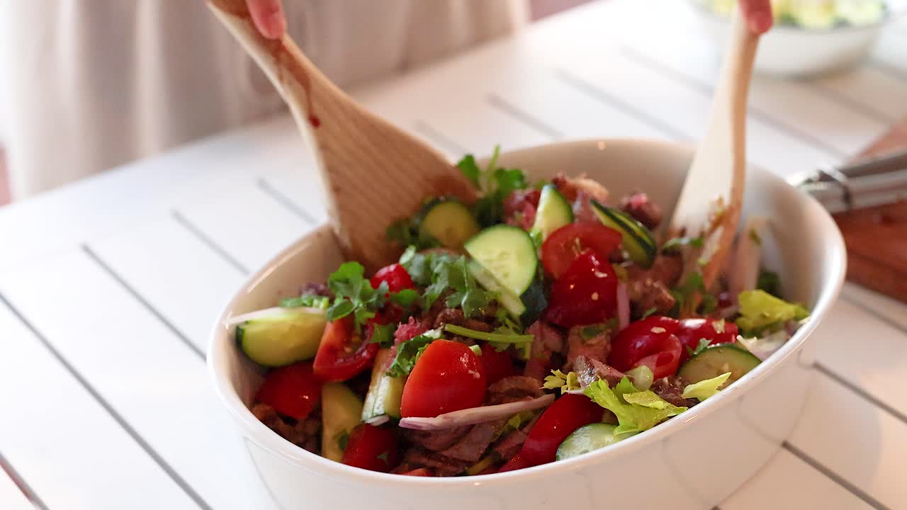 Hands mix a vibrant beef salad with cucumbers, tomatoes, and greens in a bright kitchen setting