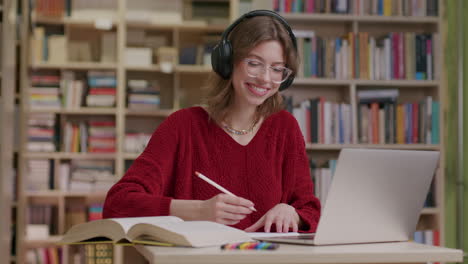 niña sonriente con gafas y auriculares estudia con una laptop en la biblioteca