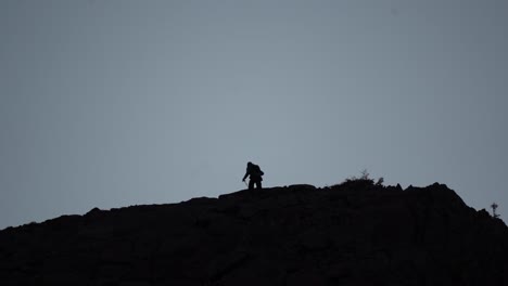 Silhouette-of-a-hiker-male-climbing-on-a-mountain-on-a-cloudy-evening,-wide-shot