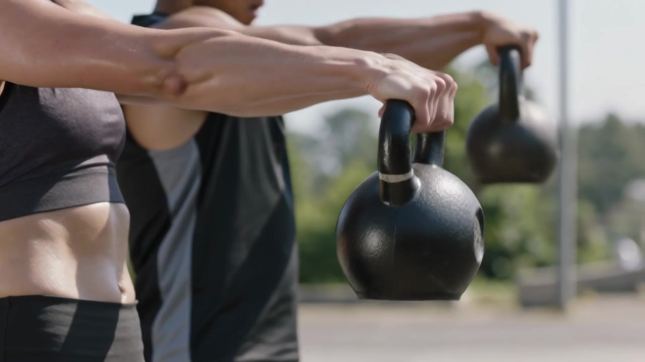 Två partners böjer sig i höfterna och lyfter kettlebells synkroniserat i en park, klädda i träningskläder, för att bygga styrka.