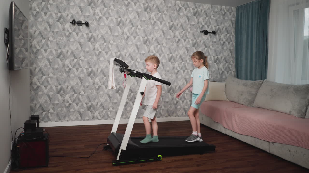 Young boy and girl walk cautiously on treadmill in living room with geometric wallpaper and soft lighting, girl stepping carefully while maintaining balance beside focused boy during indoor workout