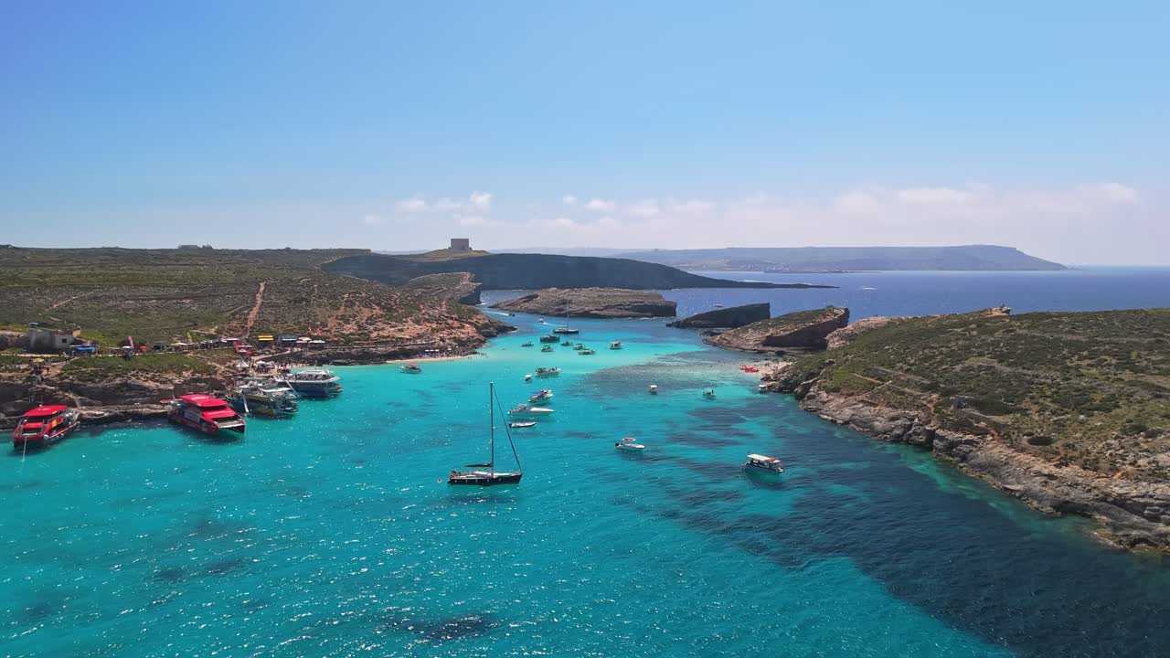 La Laguna Blu, Comino, Malta, e la vicina Cominotto sono viste dall'alto con acque turchesi, costa rocciosa, barche ancorate e una spiaggia sabbiosa sotto un cielo azzurro e limpido in una soleggiata giornata estiva.