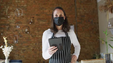 Caucasian-female-business-owner-wearing-face-mask-and-apron,-using-tablet,-looking-at-camera