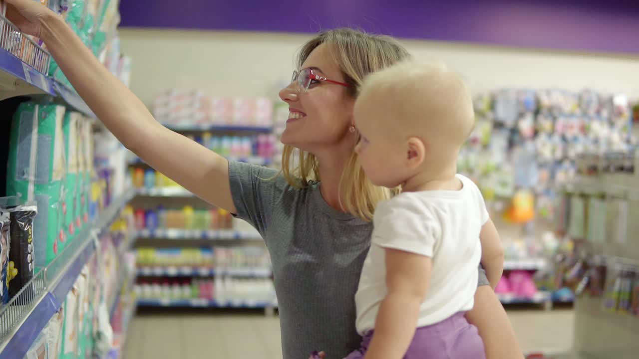joven madre atractiva con gafas sosteniendo a su hijo en sus brazos mientras elige pañales en los estantes del supermercado