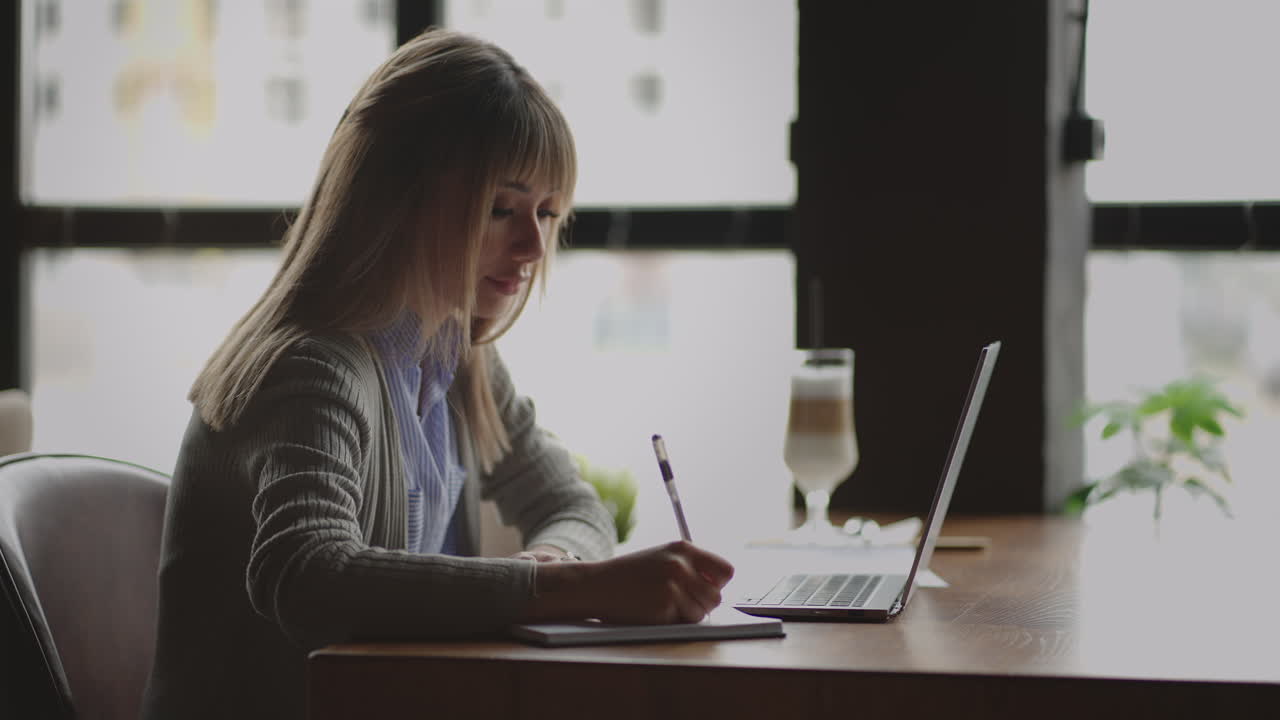 vista lateral mujer china sentada junto a la ventana con un portátil escribe en un cuaderno de casos y tareas mirando a la pantalla