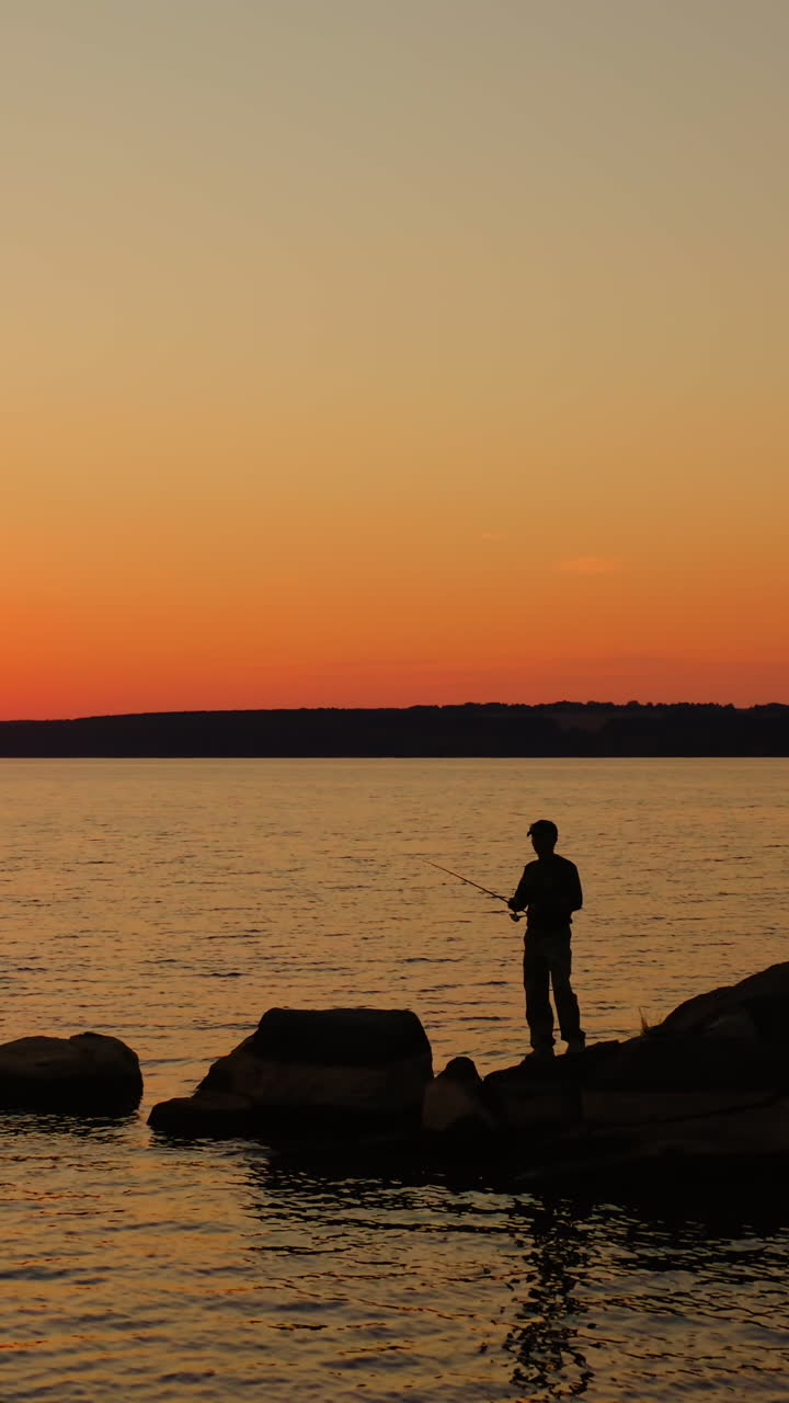 People fishing in the evening. Fishermen stand on a big stone surrounded by water at sunset. Silhouette of men with fishing rods against the setting sun. Vertical video