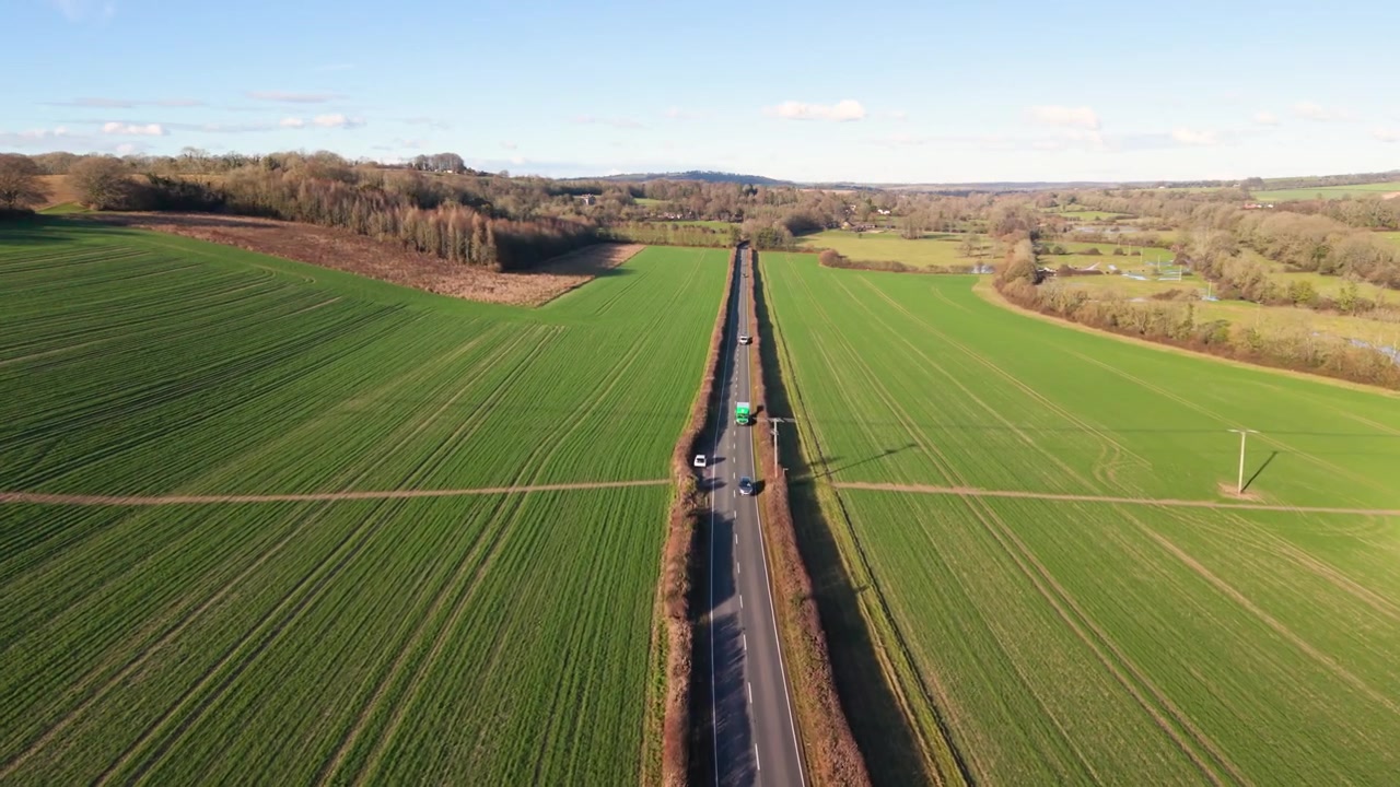 South Downs Nationalpark, langsom flyvning over landevej med passerende biler i det engelske landskab over dyrkede marker i eftermiddagssolen med udsigt til fjerne bakker og blå himmel.