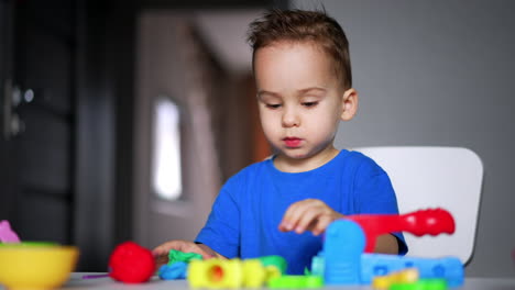 lovely baby boy in blue t-shirt focused on his game. toddler uses plasticine and plastic cups. blurred backdrop.