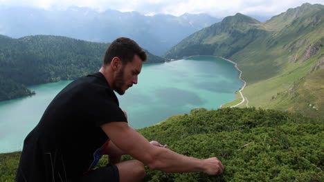 young man harvesting blueberries during a hike in the mountains, behind him lake ritom