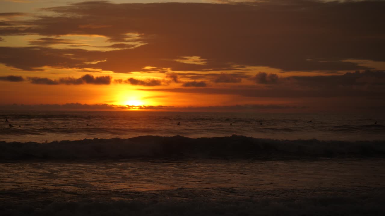 Fjerne surfere, der venter på bølger i Stillehavet under en orange solnedgang i Santa Teresa, Costa Rica. Bredt havlandskab med blide bølger, rolig atmosfære og filmisk slow motion.