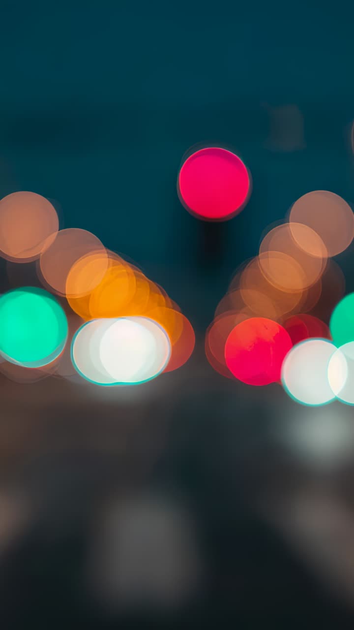 Vertical video: Pulsing red light on city street, sending amber and green bokeh with cars passing