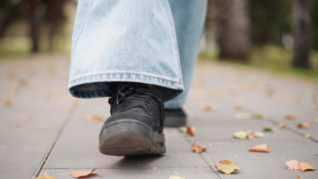 Close up of person foot in black shoe walking on gray pavement with scattered autumn leaves. subject wears light blue jeans and background shows blurred greenery and trees. Soft natural