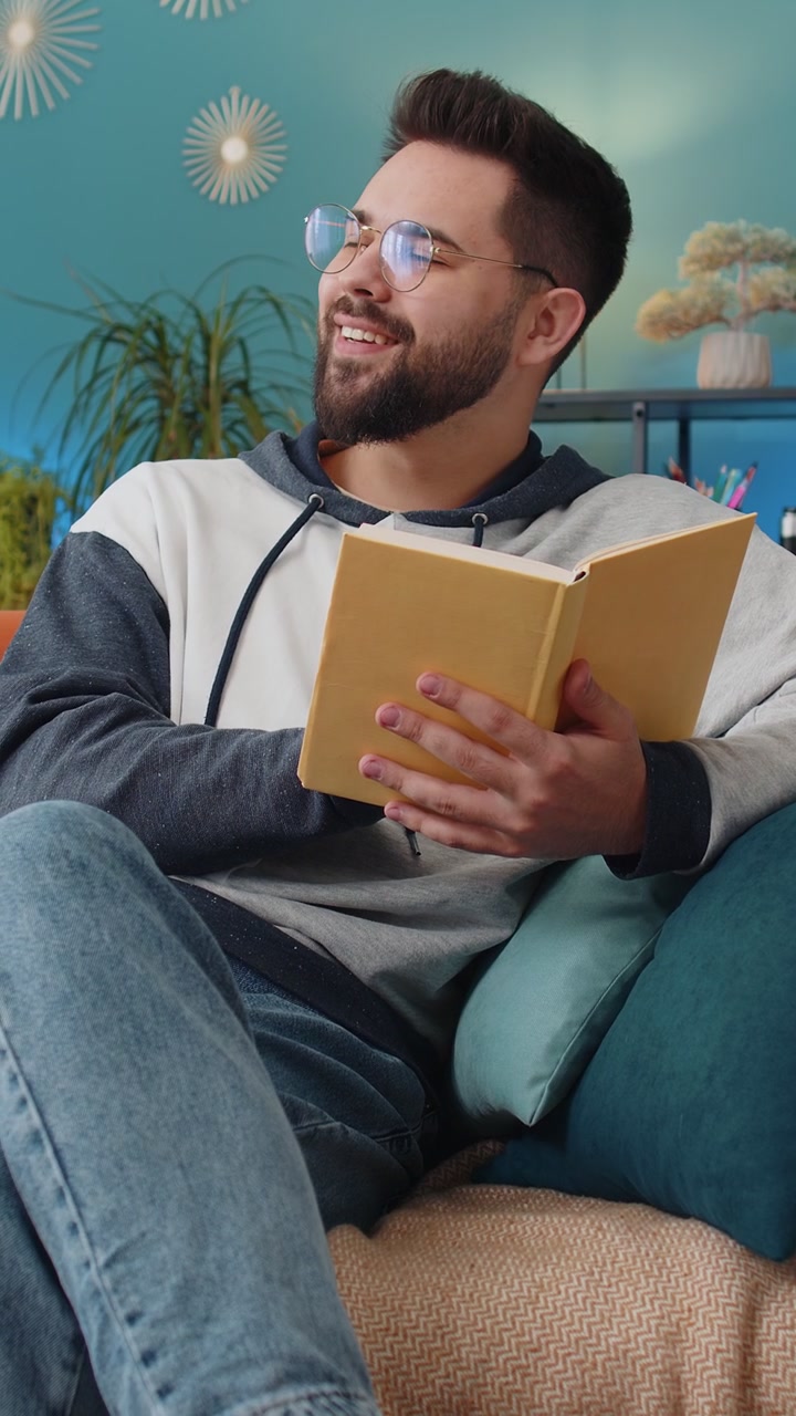 Young man relaxing interesting book turning pages smiling enjoying literature taking a rest on sofa