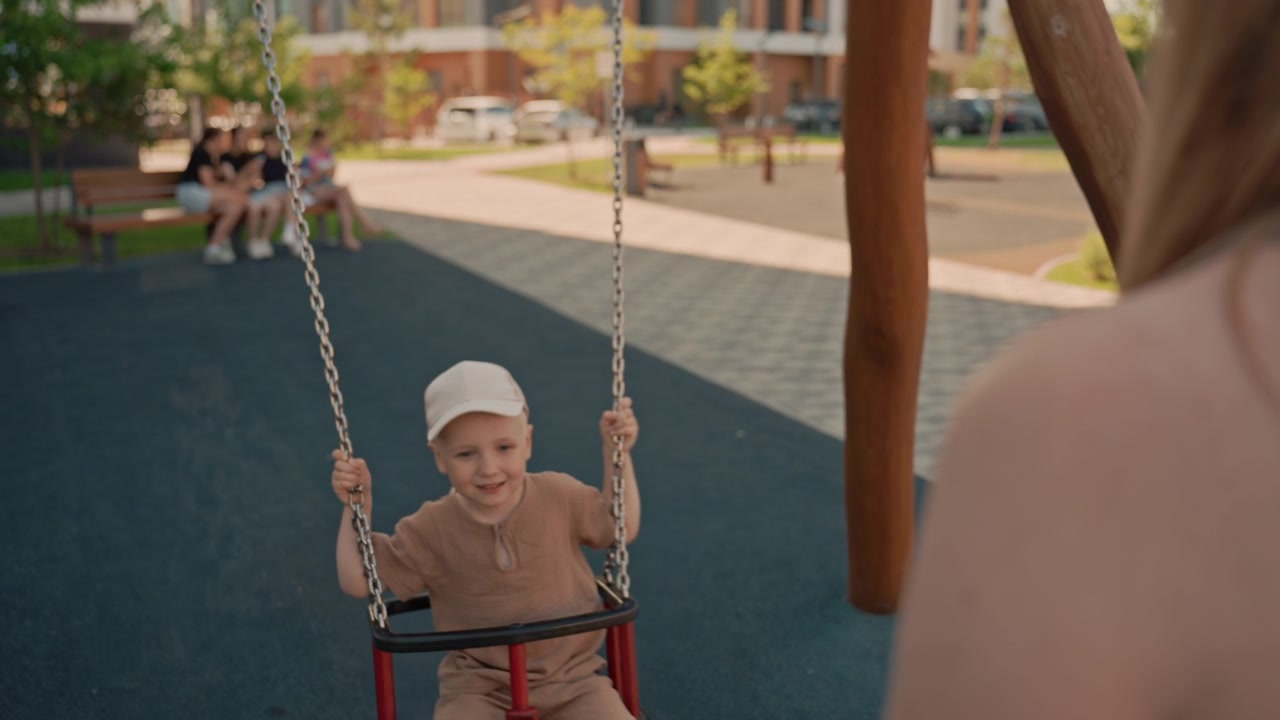 Un bambino si diverte a dondolare al parco giochi mentre una donna adulta osserva nelle vicinanze. La scena si svolge all'aperto in una giornata di sole con altre persone visibili sullo sfondo e una luce naturale soffusa che illumina