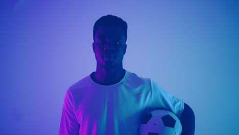 retrato en cámara lenta de un jugador de fútbol profesional negro en un uniforme blanco mirando a la cámara en la luz de neón azul-rojo del estudio. un jugador de fútbol brutal confiado y victorioso