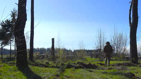 A single soldier watching a Challenger 2 4034 tank and Warrior armored vehicle driving by on a dusty gravel road from within a forest during a sunny summer day. The man is in camoflague clothing.