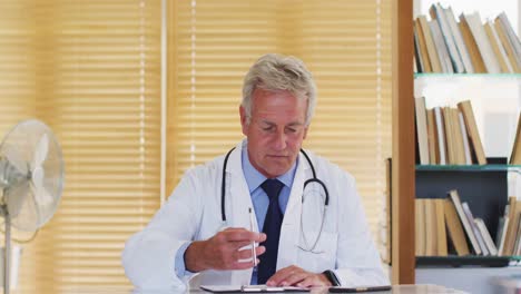 Retrato-De-Un-Médico-Caucásico-Masculino-Hablando-Y-Saludando-Mirando-A-La-Cámara-Mientras-Está-Sentado-En-El-Hospital